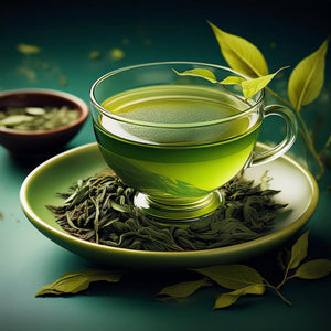 A brewed cup of green tea sits in a glass teacup on a green saucer with dry loose leaf tea on the saucer and in a dish in the background.