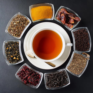 Black background with a white cup and saucer in the center with brewed tea, surrounded by glass rectangular dishes with black tea and ingredients.