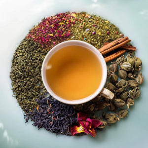 Overhead on a white background is a white teacup with brewed tea. In a circel around it is a variety of green teas and spices and herbs.