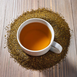 Overhead view on a light wood sits w white mug filled with tea. In a circle around is it dried and cut green rooibos.