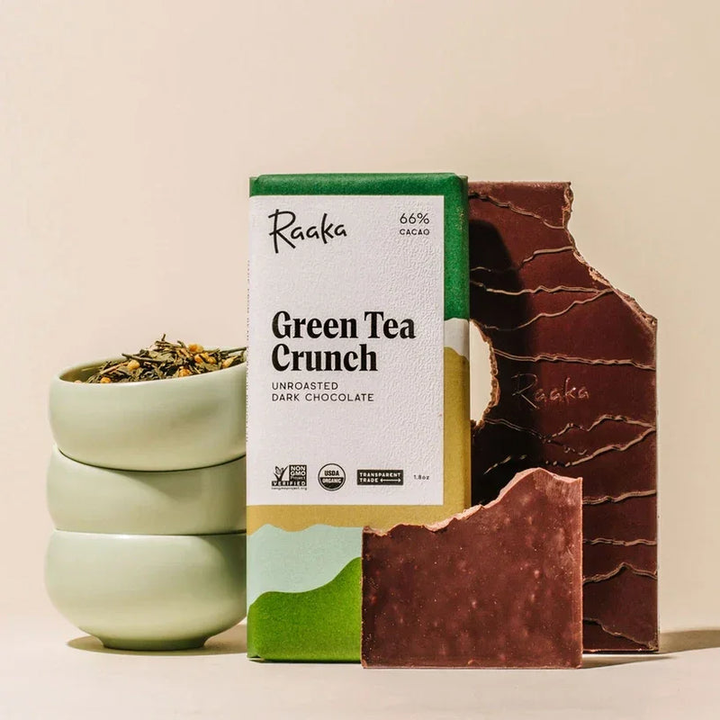 Raak Green Tea Cruch bar in the middle. On the left are a stack if green bowls with tea, on the right is the unwrapped bar with a bite taken out of it.