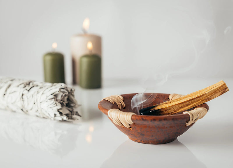 Light colored scene, In the back left corner is a trio of candles, in front of those is a  bundle of white sage. In the front right corner is a brown bowl with a partially burned palo santo stick.