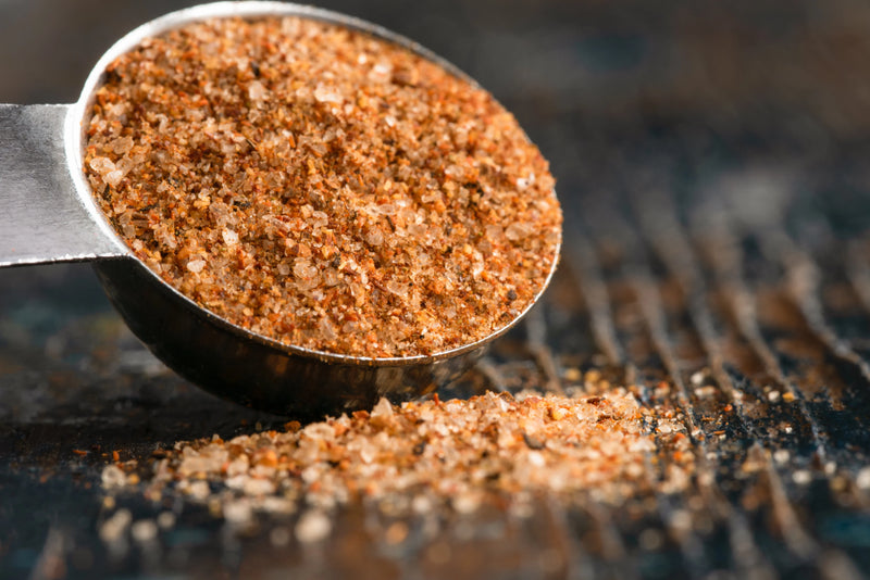 A silver measuring spoon filled with a spice blend rests on a dark surface with some of the spice blend spilled on the surface.