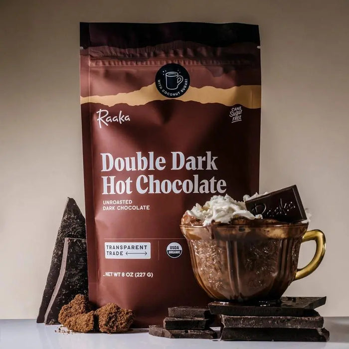 Raaka Double Dark Hot Chocolate package with a mug of hot chocolate and chocolate pieces on a neutral background