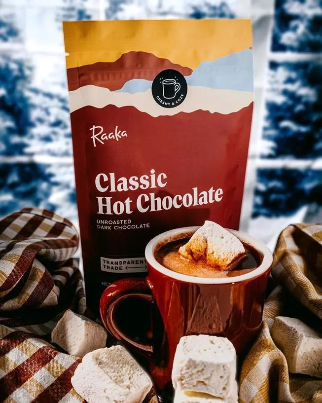 Bag of Raaka Classic Hot Chocolate with a mug of hot chocolate and marshmallows on a checkered blanket infront of a window looking out on a snow covered landscape.